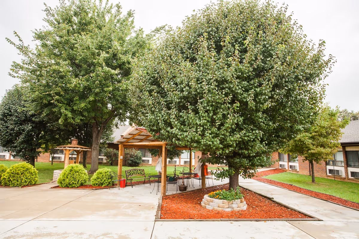 Outdoor garden area at a senior living facility with a large tree surrounded by a circular stone border filled with mulch and flowers. There is a wooden pergola with benches and chairs underneath, set on a concrete pathway. The background shows a brick building with windows and air conditioning units.
