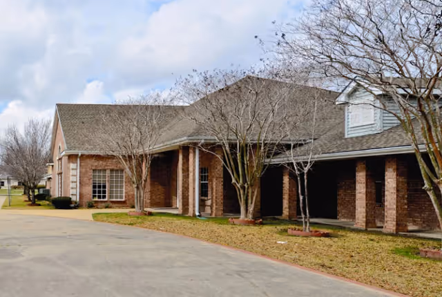 Exterior view of a single-story brick building with a sloped roof and several leafless trees in front. The building has multiple windows and a covered walkway supported by brick columns. The sky is partly cloudy.