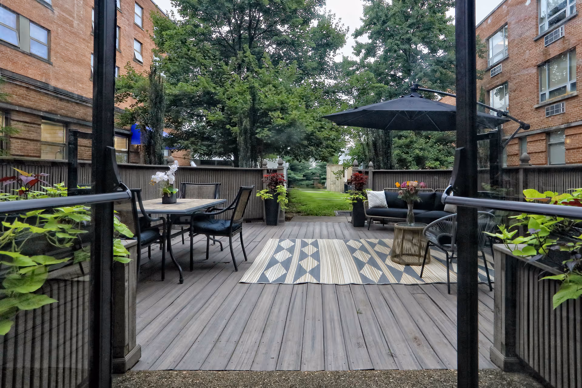 Outdoor patio area with wooden decking, featuring a dining table with chairs on the left, a seating area with a sofa, chairs, a small table, and an umbrella on the right, surrounded by plants and trees with brick buildings in the background.