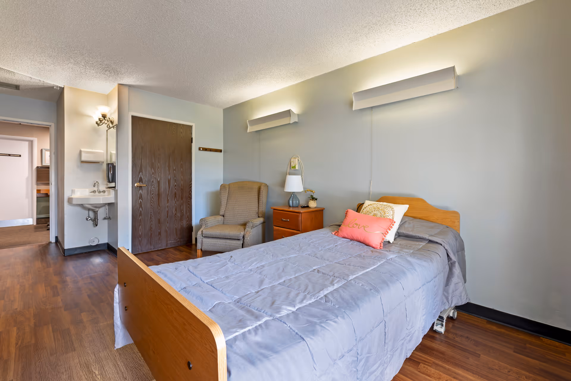 A simple bedroom in a senior living facility featuring a single bed with gray bedding and decorative pillows, a wooden nightstand with a lamp and small plant, a cushioned armchair, a wooden door, and a small sink area near the entrance. The room has wood flooring and light-colored walls with two wall-mounted light fixtures above the bed.