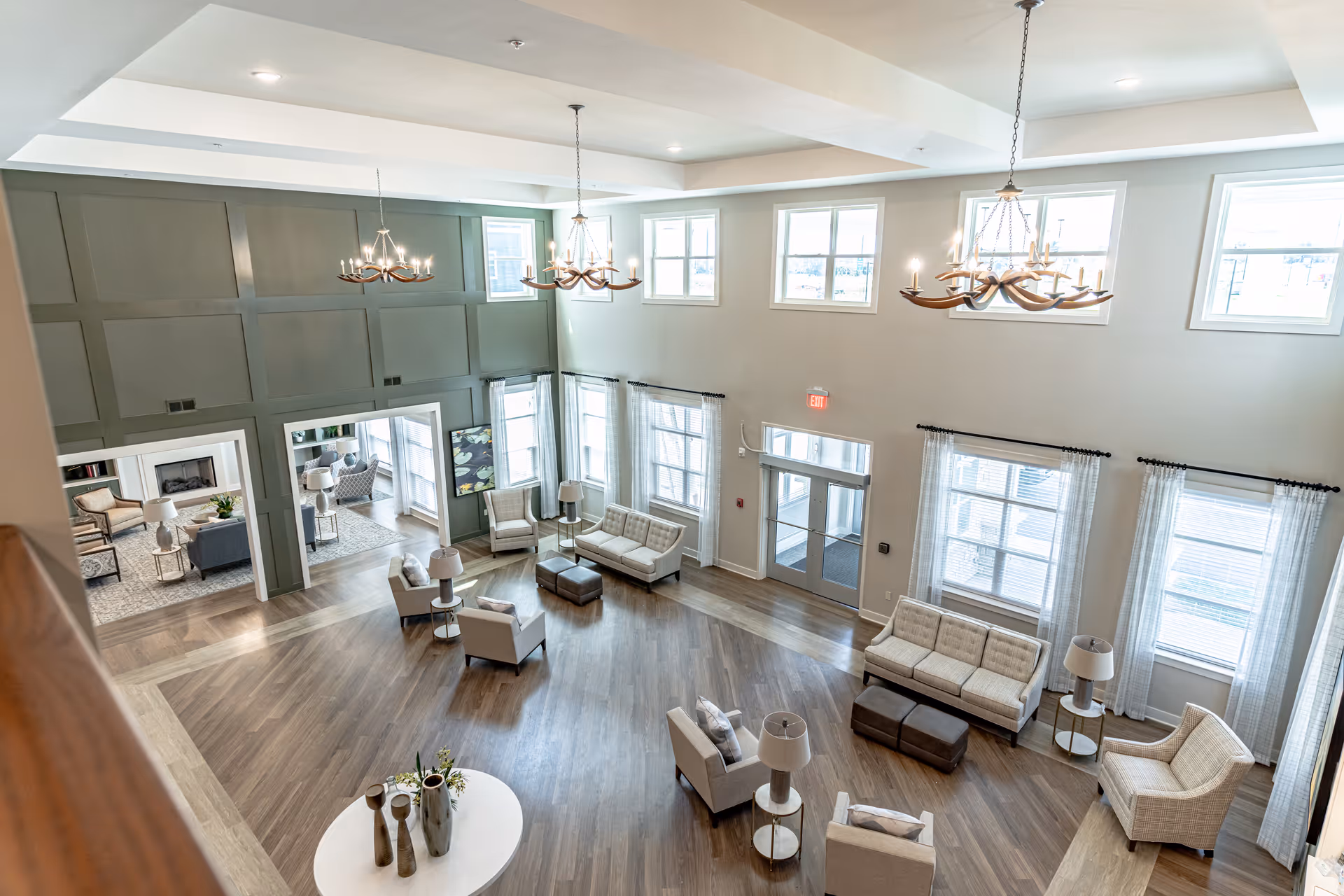 A spacious and well-lit living room area with multiple seating arrangements including sofas and armchairs, wooden flooring, large windows with sheer curtains, and three chandeliers hanging from a high ceiling. The room has a modern and cozy design with a green accent wall and an adjoining room visible through large doorways.