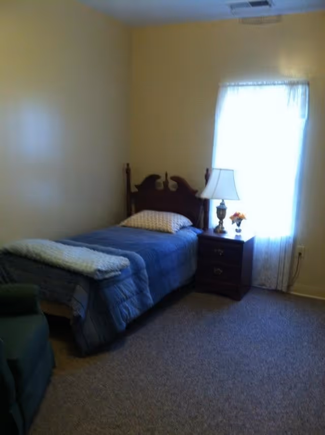 A simple bedroom with a single bed covered in a blue comforter and a pillow, a wooden nightstand with a lamp and a small flower arrangement, a window with sheer curtains letting in light, and a green upholstered chair partially visible.