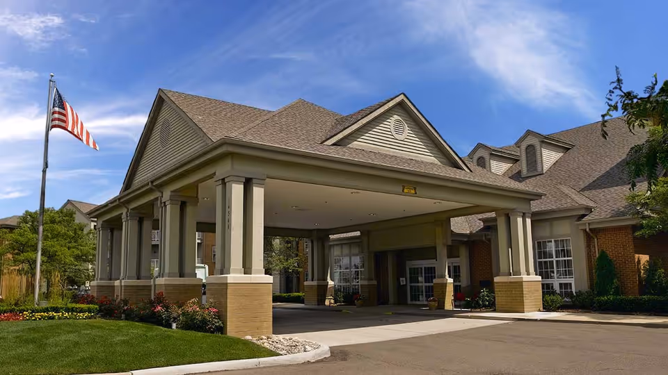 Exterior view of Shorehaven senior living facility entrance with a covered drop-off area supported by columns, an American flag on a flagpole, well-maintained landscaping, and a clear blue sky.