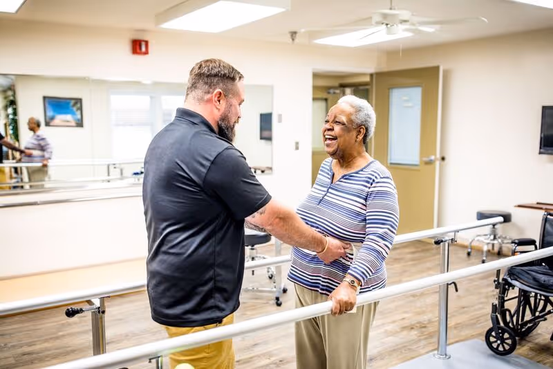 A man assisting an elderly woman with physical therapy in a rehabilitation room. The woman is holding onto parallel bars and smiling, while the man supports her by holding her waist. The room has wooden flooring, a wheelchair, a wall-mounted TV, and a large mirror on one wall.