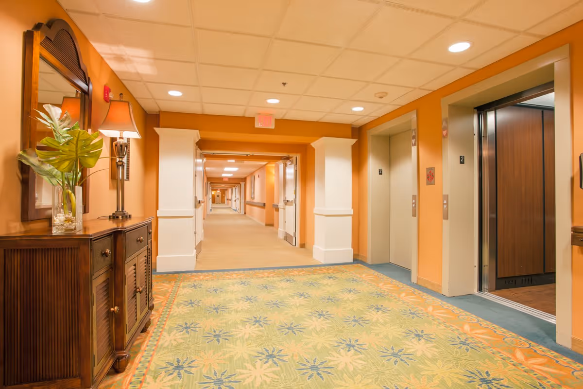 A brightly lit hallway in a senior living facility with two elevators on the right side, one elevator door open. The walls are painted orange with white trim, and there is a patterned carpet on the floor. On the left side, there is a wooden cabinet with a mirror above it, a lamp, and a vase with green leaves. The hallway extends into the distance with doors and handrails along the walls.