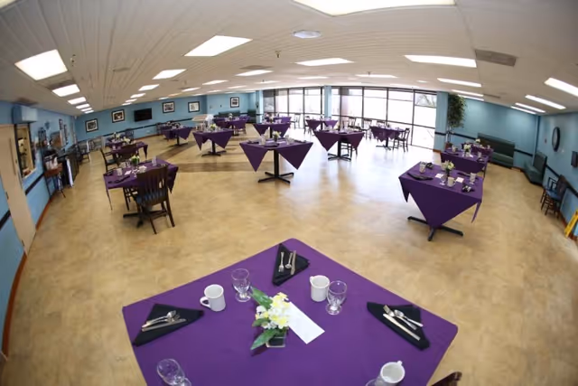 Large dining room with multiple tables covered in purple tablecloths set with dishes and silverware and windows along the far wall.