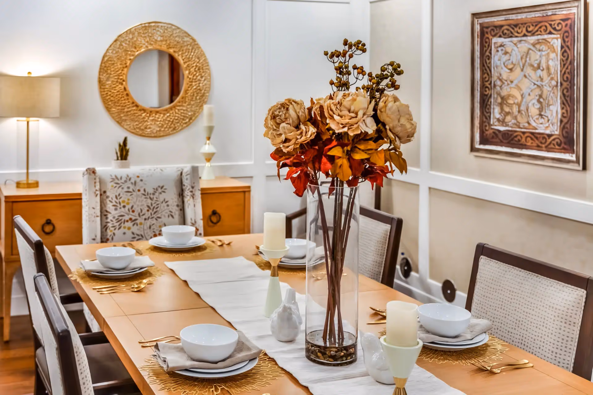 Elegantly set dining table with a tall vase of flowers, candles, place settings and chairs in a decorated dining room.