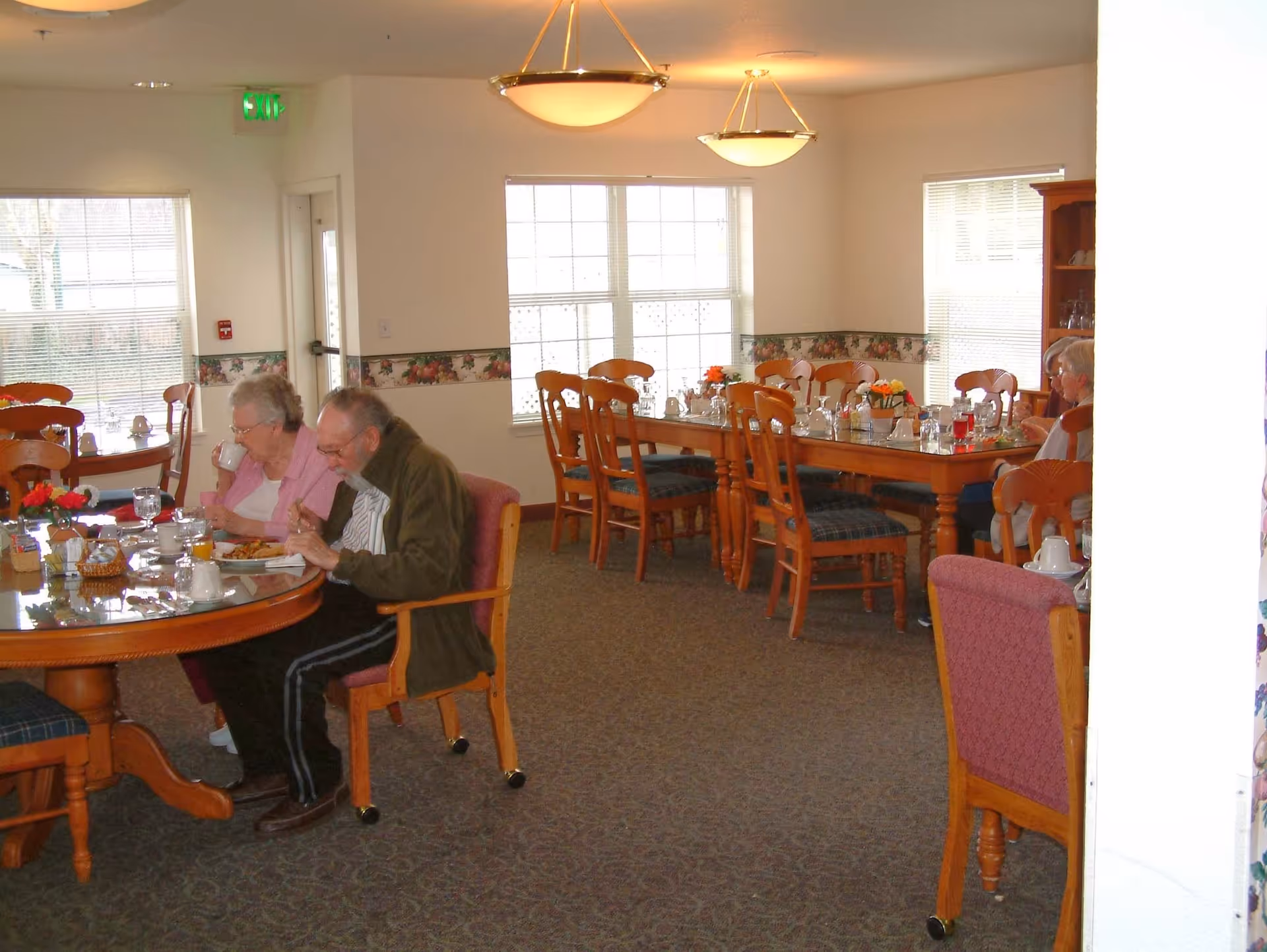 Senior living dining room with elderly residents seated at wooden tables eating a meal.
