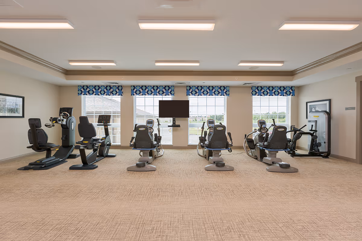 A bright fitness room with several recumbent exercise bikes and cardio machines facing a wall-mounted TV and large windows.