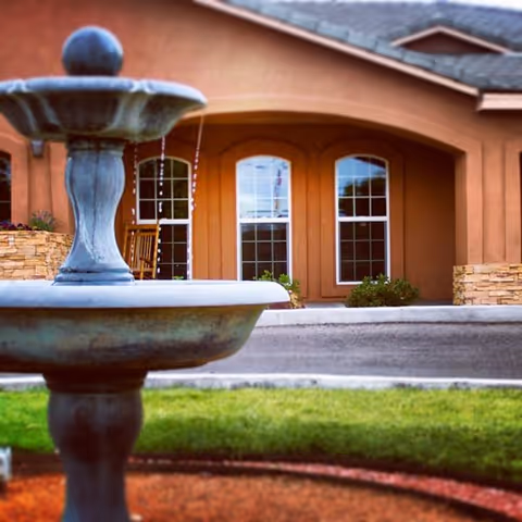 A stone fountain with flowing water in the foreground and the arched entrance and windows of a senior living building in the background.