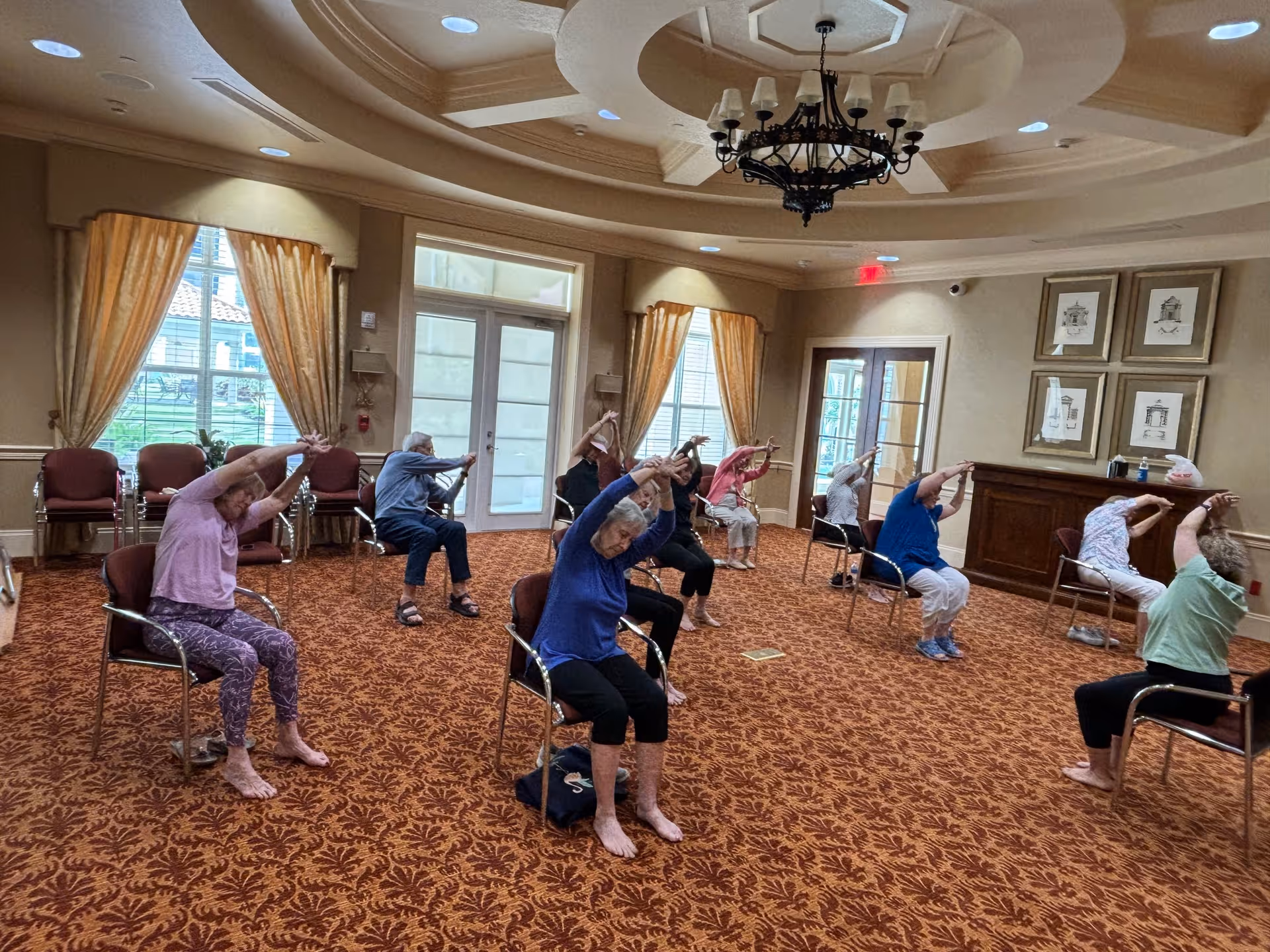 A group of elderly individuals seated on chairs in a spacious, carpeted room with large windows and elegant curtains, participating in a seated stretching exercise with their arms raised and hands clasped overhead. The room features a decorative ceiling with a chandelier and framed artwork on the walls.