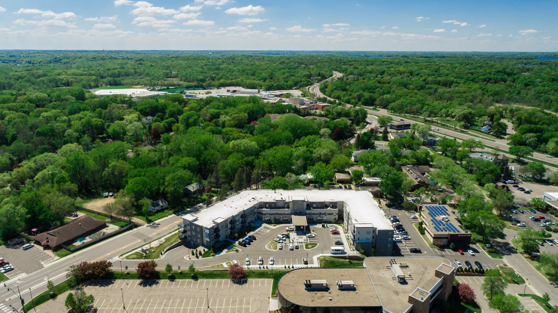 Aerial view of Havenwood of Minnetonka facility surrounded by lush green trees and roads, with parking lots and nearby buildings visible under a partly cloudy sky.