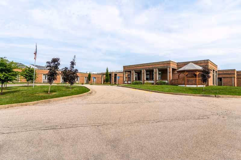 Exterior view of a single-story brick building with a driveway leading up to the entrance. The building has a covered porch area and a wooden gazebo on the right side. There are several small trees and a flagpole with an American flag on the left side, set against a partly cloudy sky.