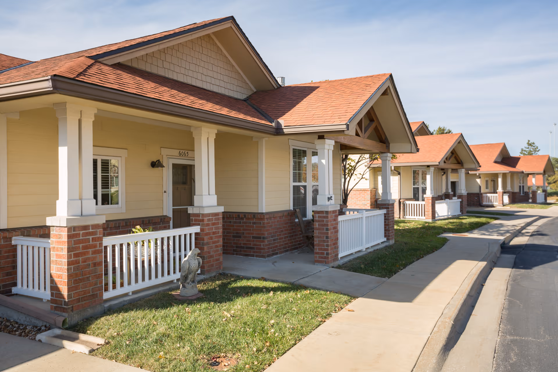 Exterior view of a row of single-story senior living cottages with beige siding, red brick accents, white railings, and red shingled roofs under a clear sky.