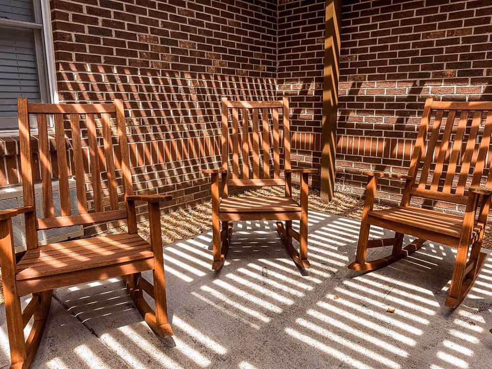 Three wooden rocking chairs placed on a concrete patio with shadows of a pergola creating striped patterns on the chairs and ground, against a brick wall background.