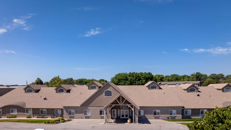 Front exterior of a single-story assisted living building with a peaked entrance canopy, multiple dormer windows, and a clear blue sky.