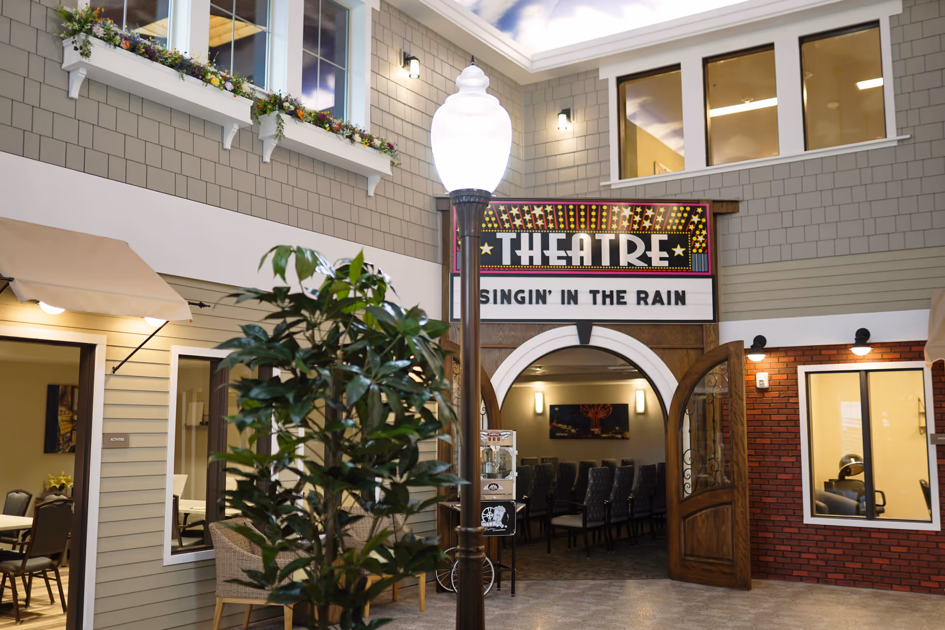 Indoor area designed to look like a small town street with a theatre entrance displaying a sign for 'Singin' in the Rain'. There are windows with flower boxes above, a street lamp in the center, and seating visible inside the theatre. The walls have a mix of siding and brick finishes, and there is a popcorn machine near the theatre entrance.
