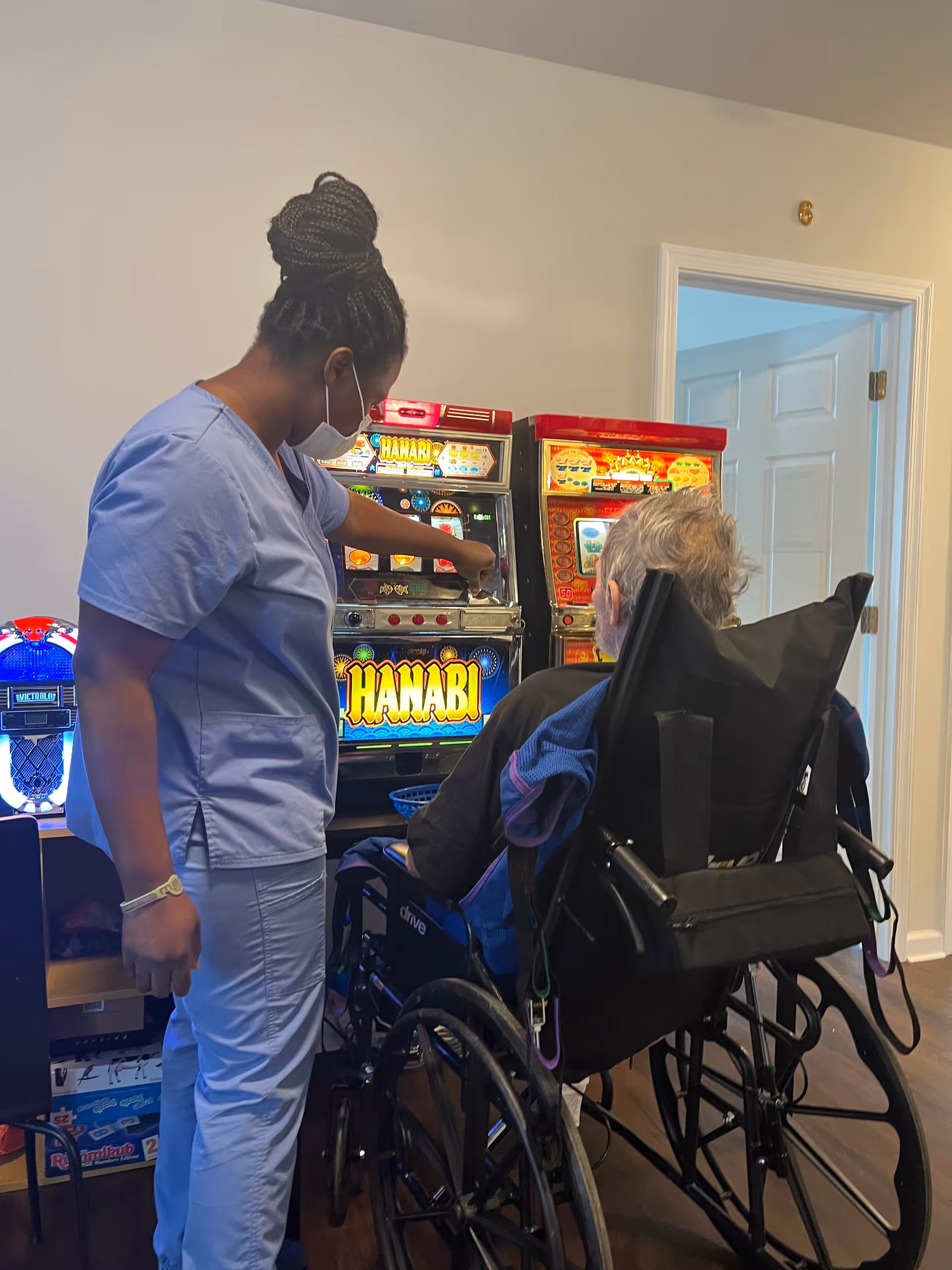 A caregiver wearing scrubs and a face mask assists an elderly person in a wheelchair playing on a slot machine in an indoor recreational area.