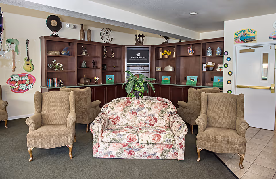A cozy living room area with a floral patterned loveseat centered between two brown upholstered armchairs. Behind the seating is a large wooden built-in bookshelf filled with decorative items, books, and plants. The wall has musical-themed decorations including guitars and a 'Rock & Roll' sign. A door with a 'Surfin' USA' sign and vinyl record decorations is visible on the right.