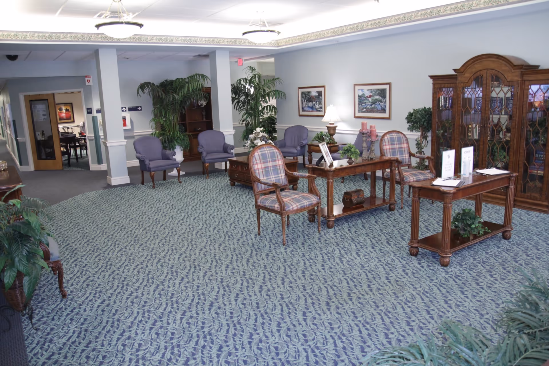 A spacious, well-lit common area with patterned carpet, several upholstered chairs, wooden tables, decorative plants, framed artwork on the walls, and a large wooden cabinet with glass doors.