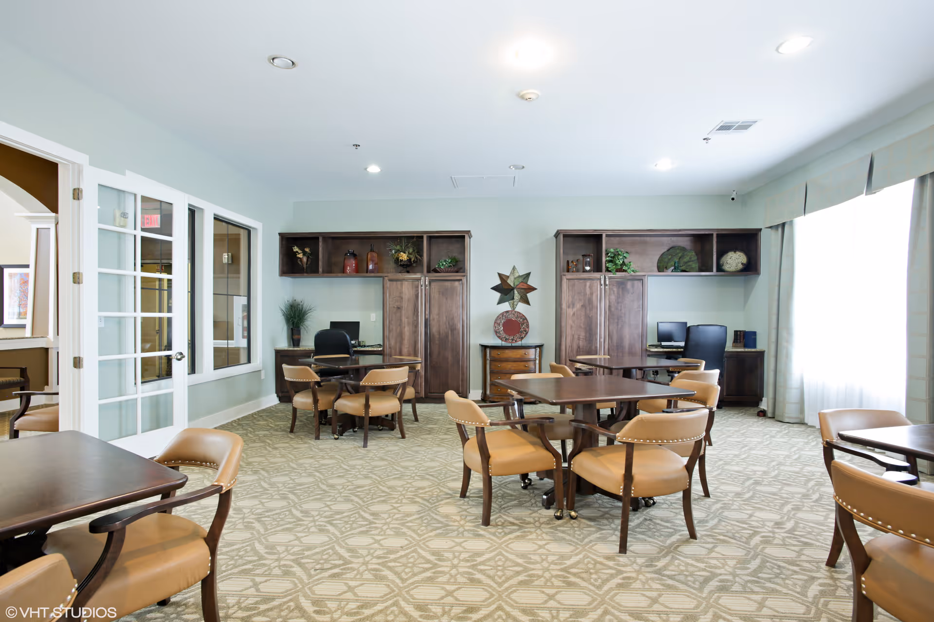 A well-lit common area with multiple wooden tables and tan cushioned chairs arranged on a patterned carpet. The room features large windows with curtains on the right side, two wooden cabinets with shelves holding decorative items, and two desks with office chairs and computers. There is a glass door on the left side leading to another room.