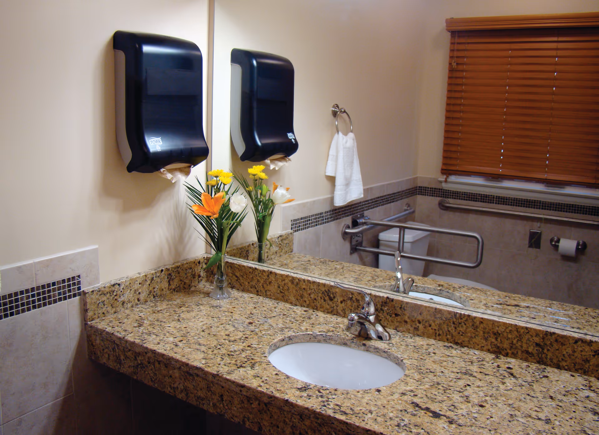 A clean bathroom with a granite countertop and an oval sink. There is a large mirror above the sink, a black paper towel dispenser mounted on the wall, a vase with orange and yellow flowers on the counter, a white hand towel hanging on a ring, and a window with wooden blinds. The bathroom also features grab bars near the toilet for accessibility.