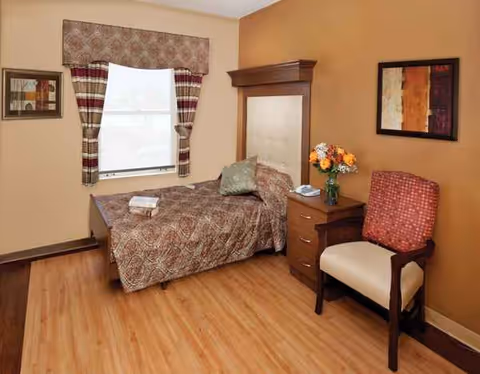 A furnished bedroom in a senior facility featuring bed, nightstand, chair, and window with curtains.