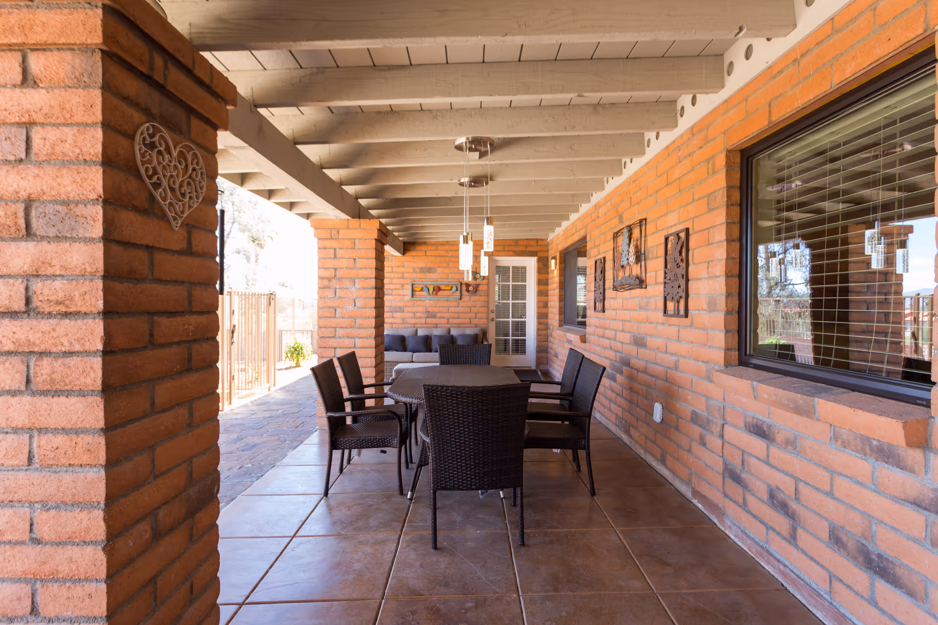 Covered outdoor patio area with brick walls and tiled floor, featuring a rectangular table with six wicker chairs and a cushioned sofa against the far wall. Decorative wall art and hanging pendant lights are visible, along with a window with blinds on the right side.