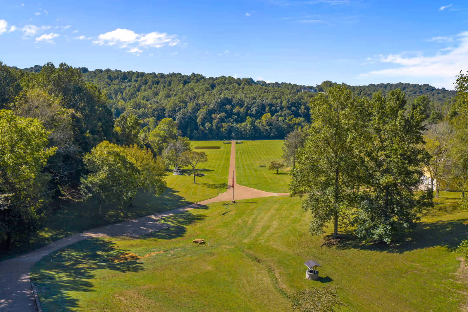 A wide, green outdoor area with a long, straight pathway running through the center, surrounded by trees and grassy fields under a blue sky with scattered clouds.