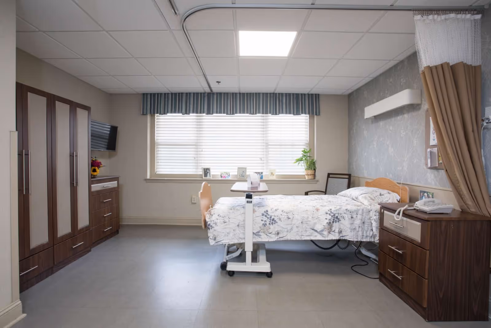 A clean and well-lit senior living bedroom with a hospital-style bed covered in a floral bedspread. The room features a large window with blinds and a striped valance, a wooden dresser with a telephone, a chair, a small table on wheels over the bed, and a wardrobe with multiple doors. A privacy curtain is partially drawn on the right side of the room.