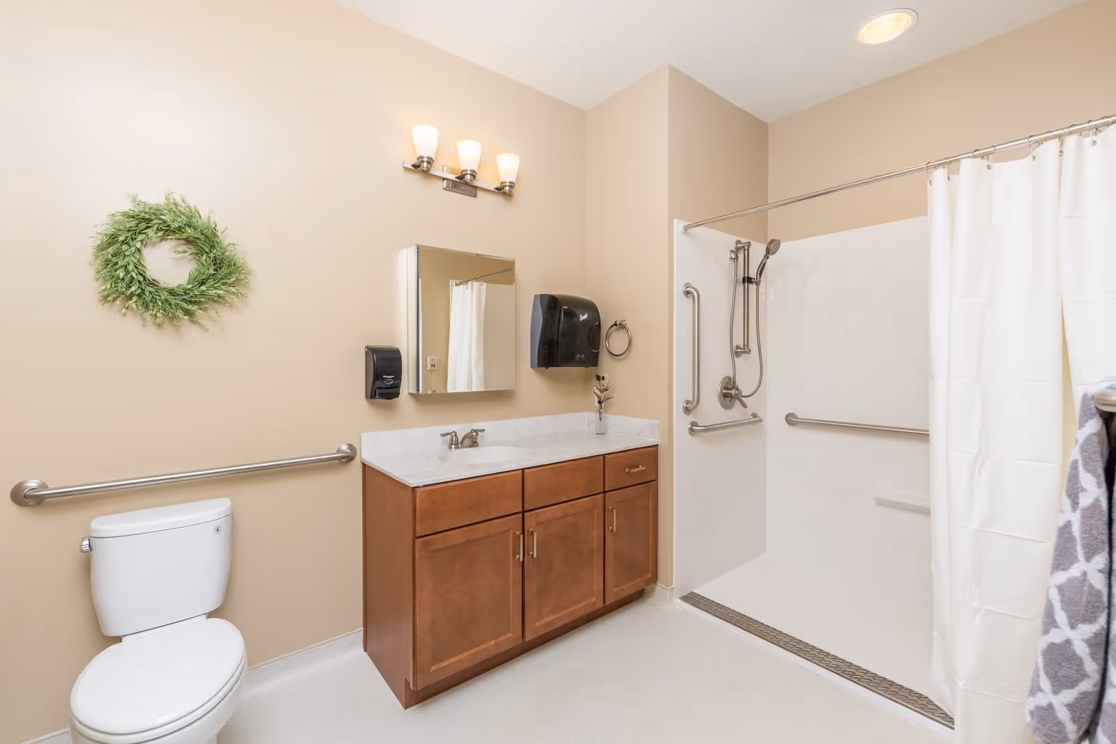 Accessible bathroom with a toilet, wooden vanity and mirror, and a roll-in shower with grab bars and a curtain.