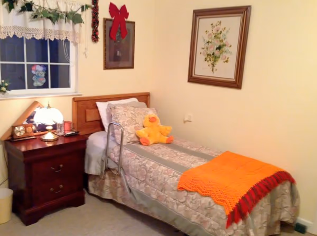 A cozy single bed with floral bedding and an orange throw blanket in a senior living facility room. A yellow stuffed duck sits on the bed. Next to the bed is a wooden nightstand with a lamp, clock, and other small items. The room has cream-colored walls with framed floral artwork and a window with lace curtains and green garland.
