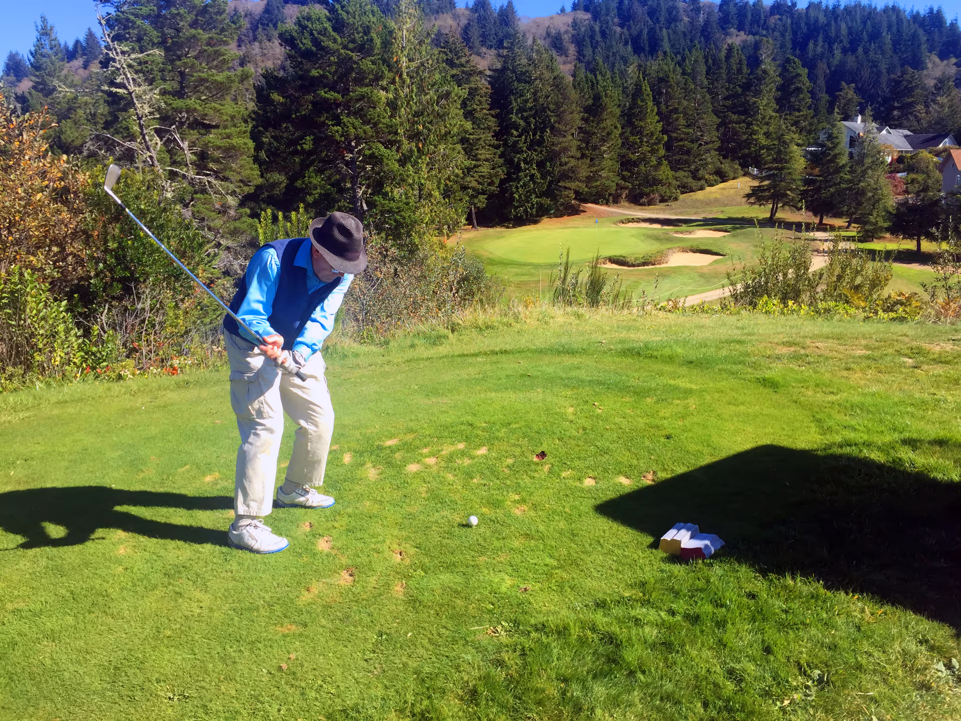 An elderly man wearing a hat, blue shirt, and beige pants is playing golf on a green golf course surrounded by trees and hills under a clear blue sky.