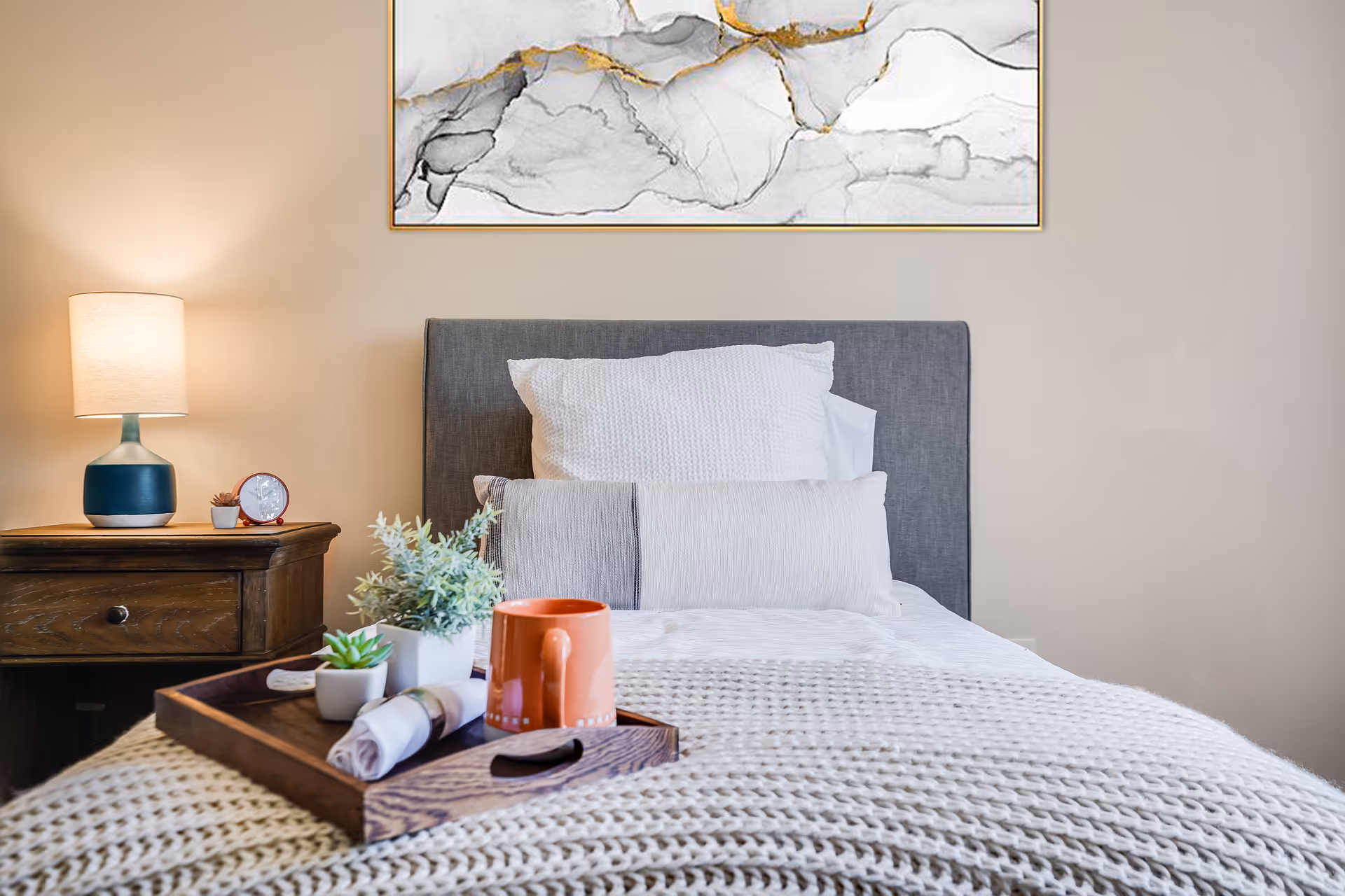 Neatly made bed with a gray headboard, a wooden tray holding a mug and small plants, a bedside table with a lamp and clock, and artwork above.