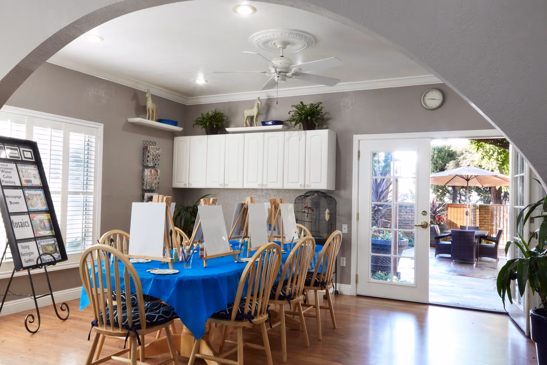 A bright and inviting activity room with a large table covered in a blue tablecloth, surrounded by wooden chairs with cushions. The table is set up with easels, blank canvases, and painting supplies. The room has light gray walls, white cabinets, a ceiling fan, and a large window with white shutters. French doors open to an outdoor patio area with seating and an umbrella. There are plants and decorative items on shelves and a display board listing various art activities.