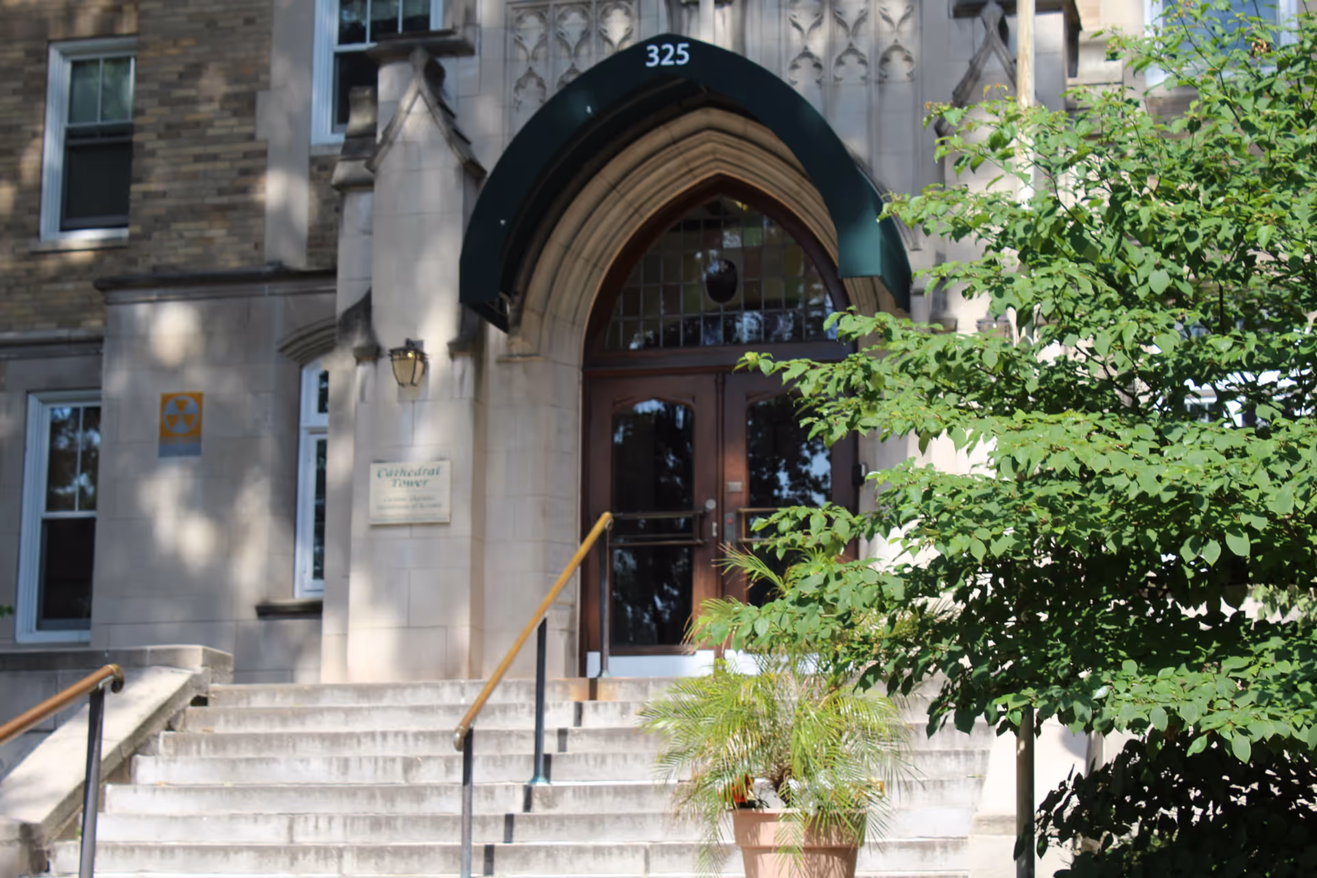Stone building entrance with a green awning displaying the number 325 above double wooden doors. Stone steps with handrails lead up to the entrance, and there is a potted plant and a leafy tree on the right side. A plaque on the wall reads 'Cathedral Tower'.
