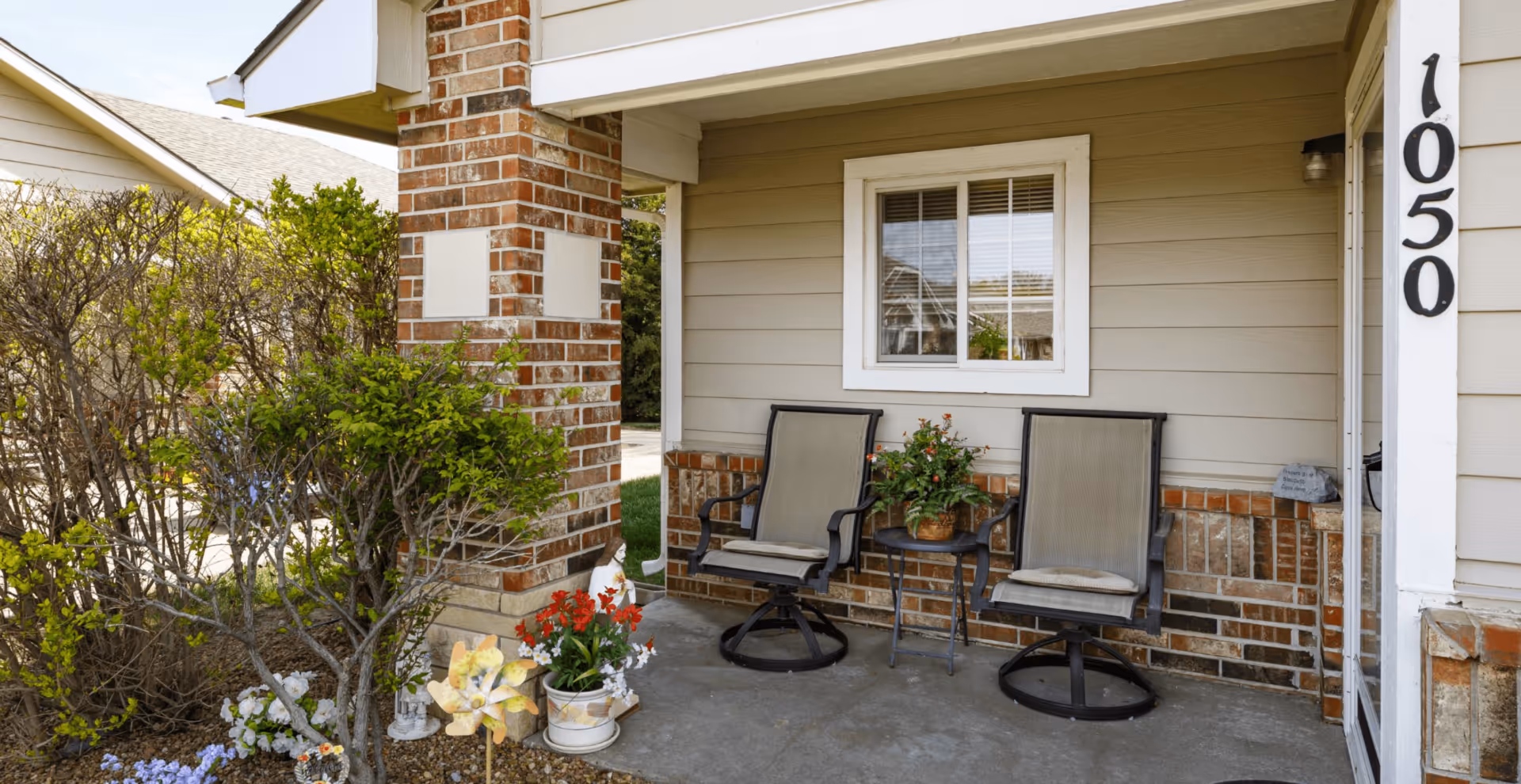 Covered front porch with two outdoor chairs, potted plants, a brick column, and house number 1050.
