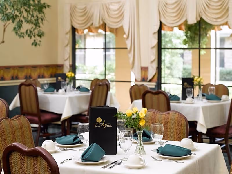A dining room with tables covered in white tablecloths, each set with green folded napkins, glassware, plates, and silverware. One table has a black menu with the word 'Atria' and a tree logo on it, along with a small vase of yellow flowers. The room has large windows with cream-colored drapes and upholstered chairs around the tables.