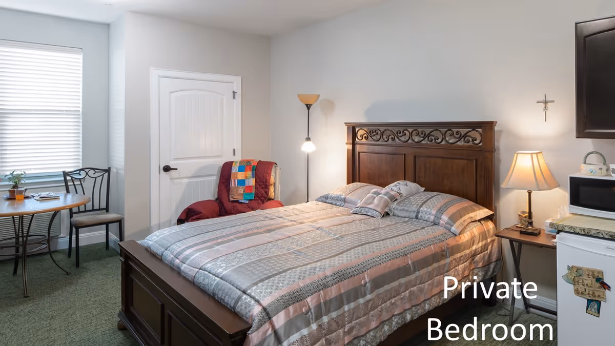 A cozy private bedroom with a large wooden bed featuring a patterned bedspread and pillows. There is a floor lamp and a bedside table with a lamp and a microwave on top. A small refrigerator is next to the bedside table. A round table with two chairs is near a window with blinds. A colorful quilt is draped over a red armchair next to a closed white door.