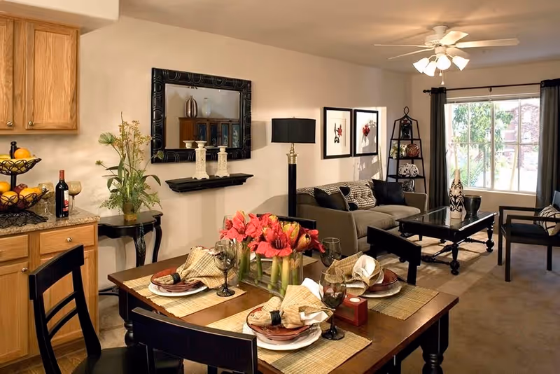 A cozy and well-decorated living and dining area in a senior living facility. The dining table is set with plates, napkins, wine glasses, and a centerpiece of red flowers. The living room features a gray sofa with cushions, a black coffee table, a floor lamp, framed artwork on the wall, and a window with curtains letting in natural light. Wooden kitchen cabinets and a fruit basket are visible on the left side.