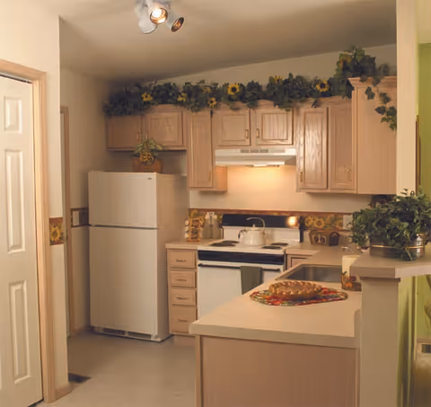 A small kitchen with light wood cabinets, a white refrigerator, a white stove with a kettle on it, and a countertop with a pie on a colorful placemat. There are decorative plants and flowers above the cabinets and on the counter.