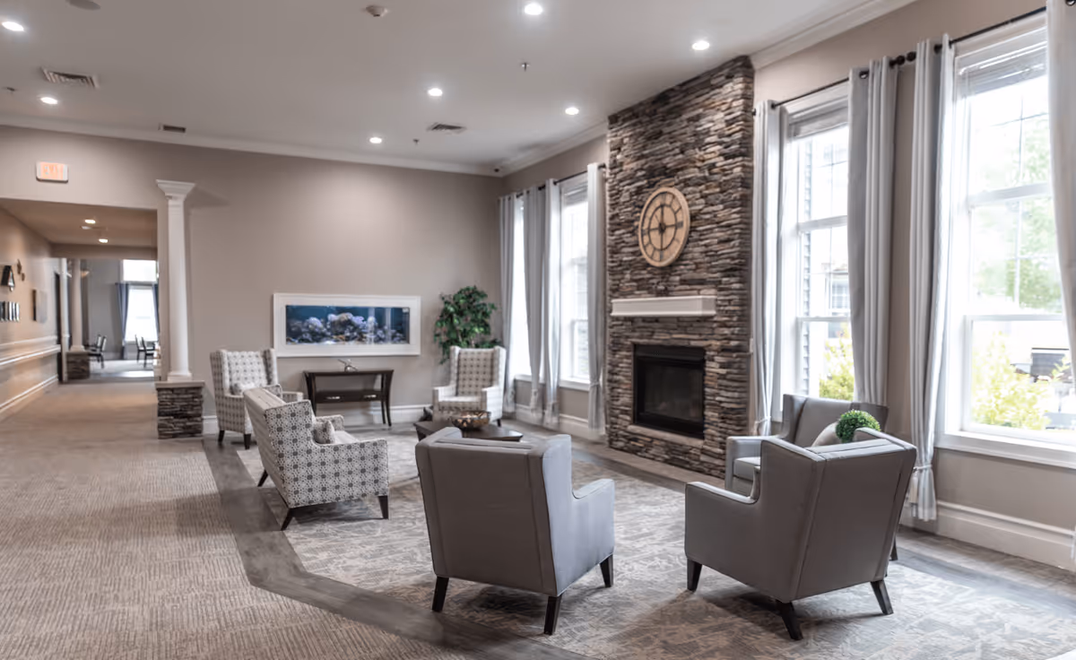 A cozy living room area in a senior living facility with four gray armchairs and two patterned armchairs arranged around a coffee table on a patterned rug. The room features a stone fireplace with a large clock above it, large windows with gray curtains letting in natural light, and a wall-mounted aquarium. The walls are painted beige, and there is a hallway visible in the background.