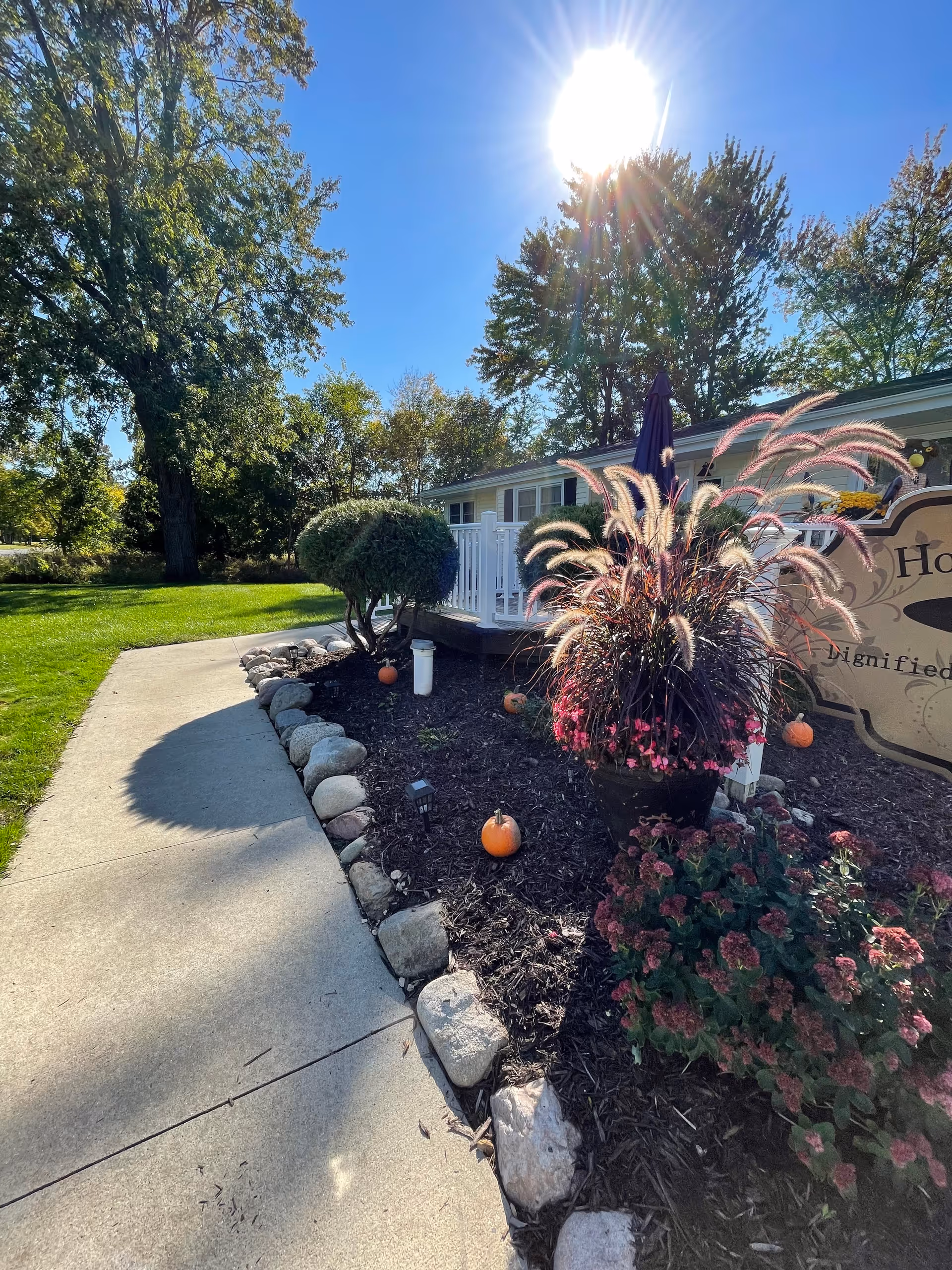 Sunlit walkway and landscaped entrance with pumpkins, ornamental grasses, flowers and a partial sign in front of a single-story senior living building.