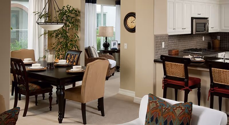 A cozy and well-lit interior space featuring a dining area with a dark wooden table set for four, beige upholstered chairs, and a hanging light fixture above. Adjacent to the dining area is a kitchen with white cabinets, a tiled backsplash, and a counter with two bar stools. In the background, there is a living area with a wicker chair, a side table with a lamp, and a large green plant near a window with white curtains.