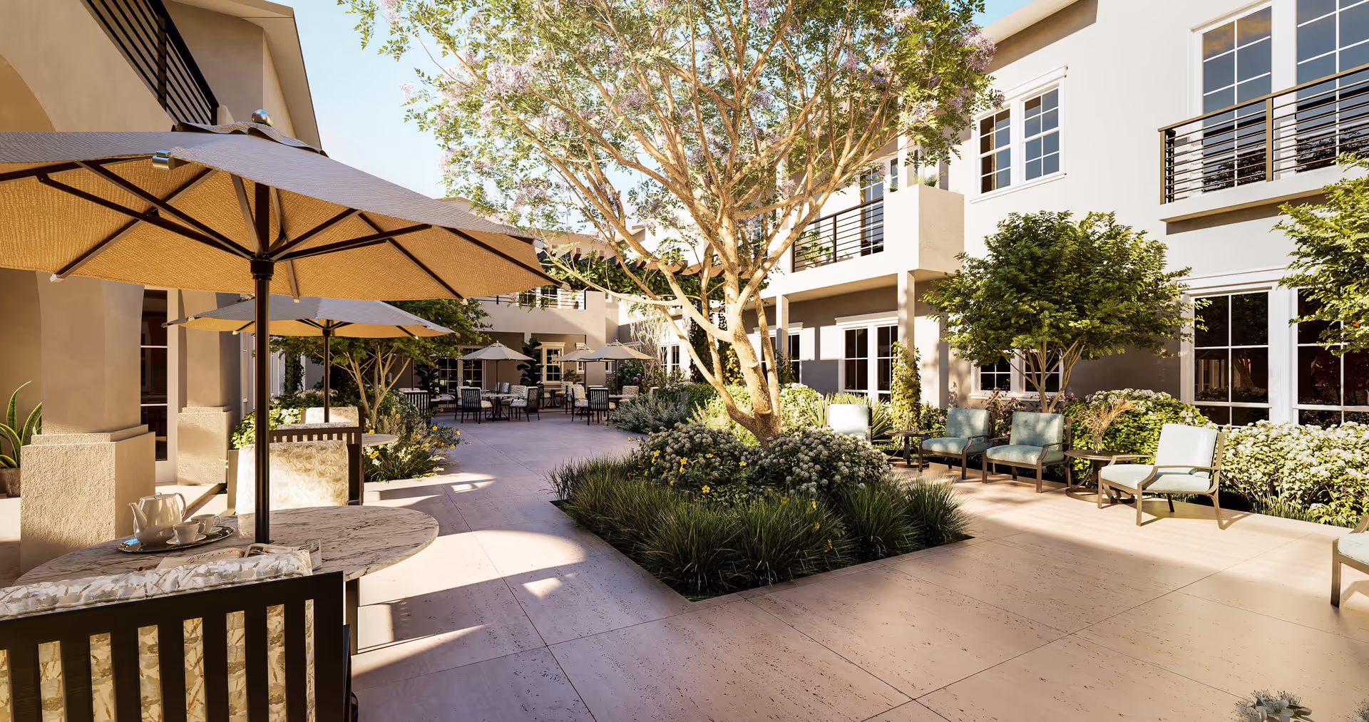 Outdoor courtyard area at Cogir of Brea featuring patio tables with umbrellas, chairs, landscaped greenery, and a large tree in the center, surrounded by a two-story building with windows and balconies.