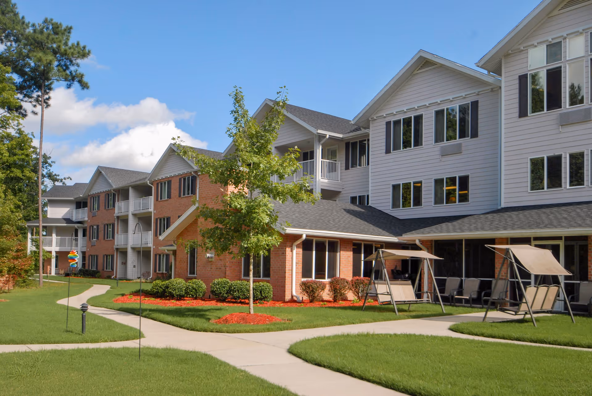 Exterior view of a multi-story senior living facility with brick and white siding. The building is surrounded by well-maintained green lawns, small trees, and landscaped flower beds with red mulch. There are concrete walkways and outdoor seating areas with swing benches under canopies. The sky is blue with some clouds.