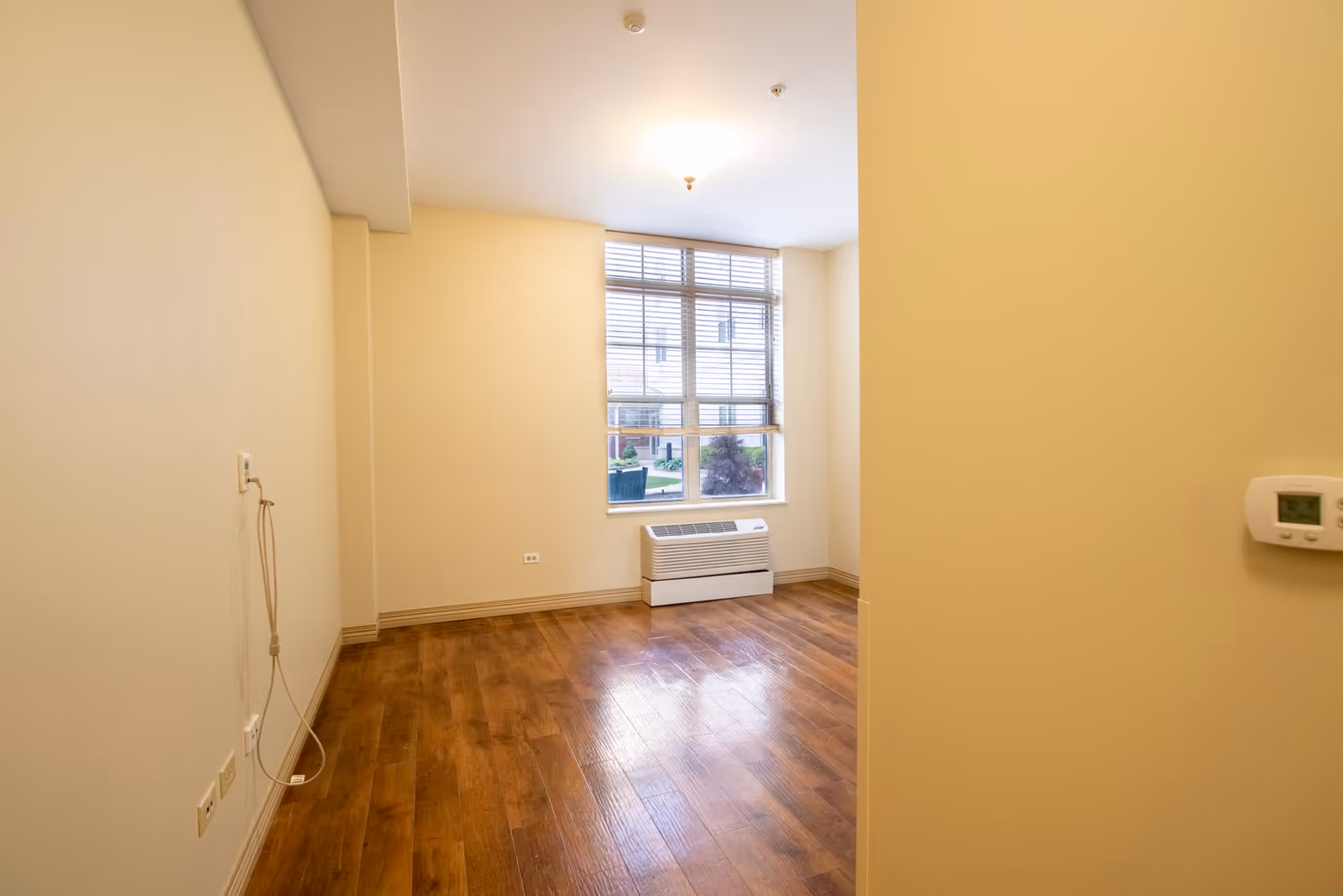 Empty room with wooden flooring, beige walls, a large window with blinds, a wall-mounted thermostat, and a heating/cooling unit below the window.