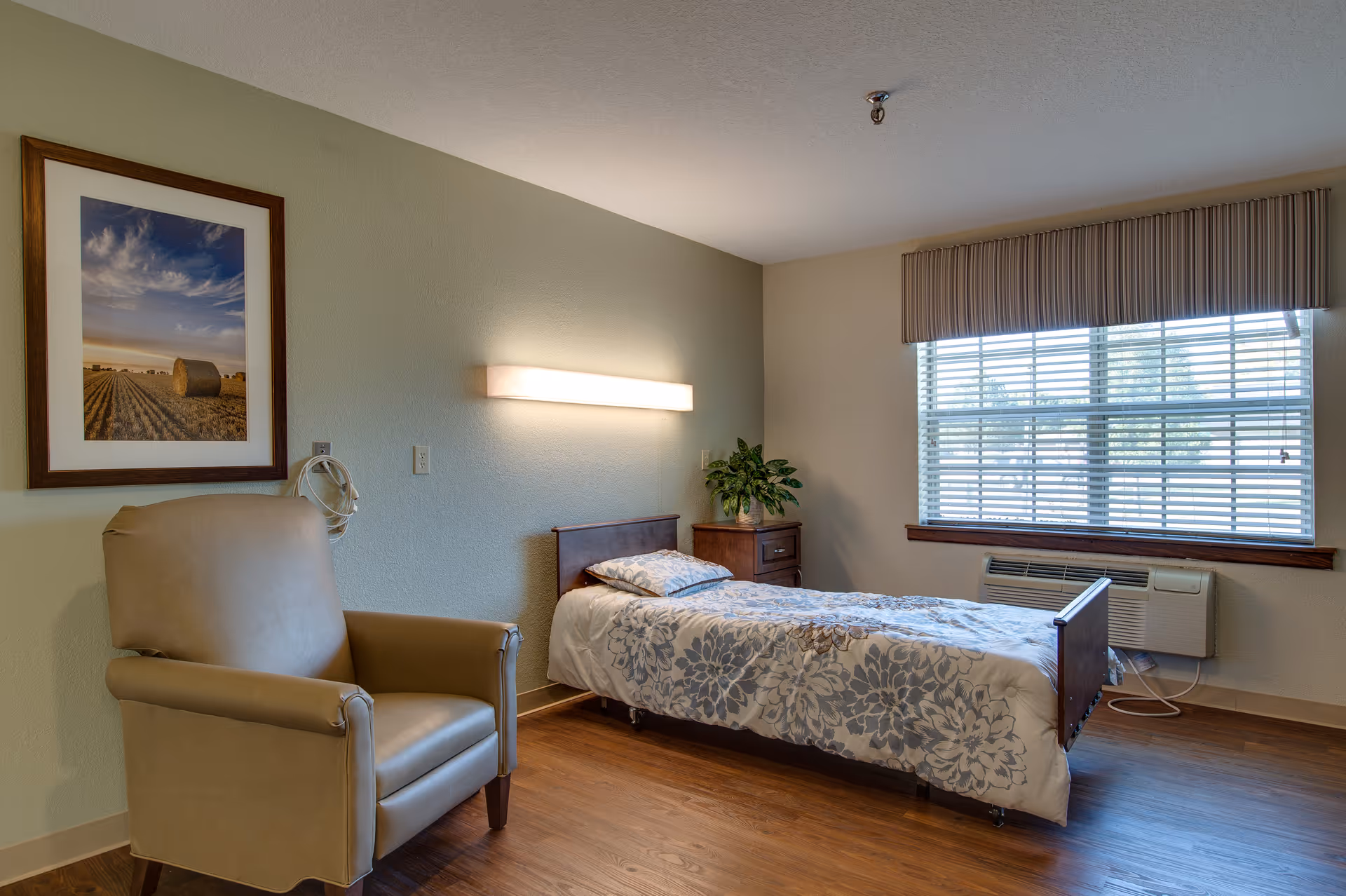 A simple, clean bedroom in a senior living facility with a single bed covered in a floral patterned comforter, a beige armchair, a wooden nightstand with a plant, a large window with blinds, and a framed landscape photograph on the wall.