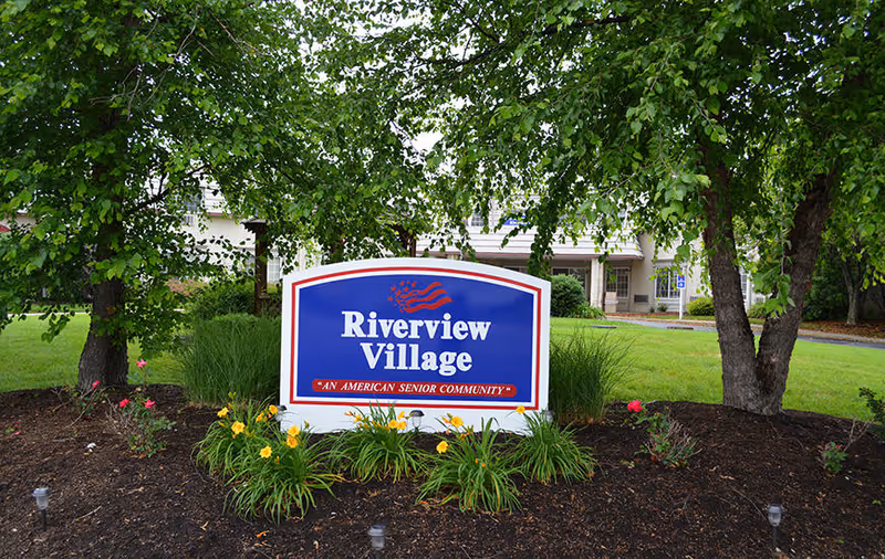 Entrance sign reading "Riverview Village" in a landscaped flower bed with trees and the facility building visible behind it.