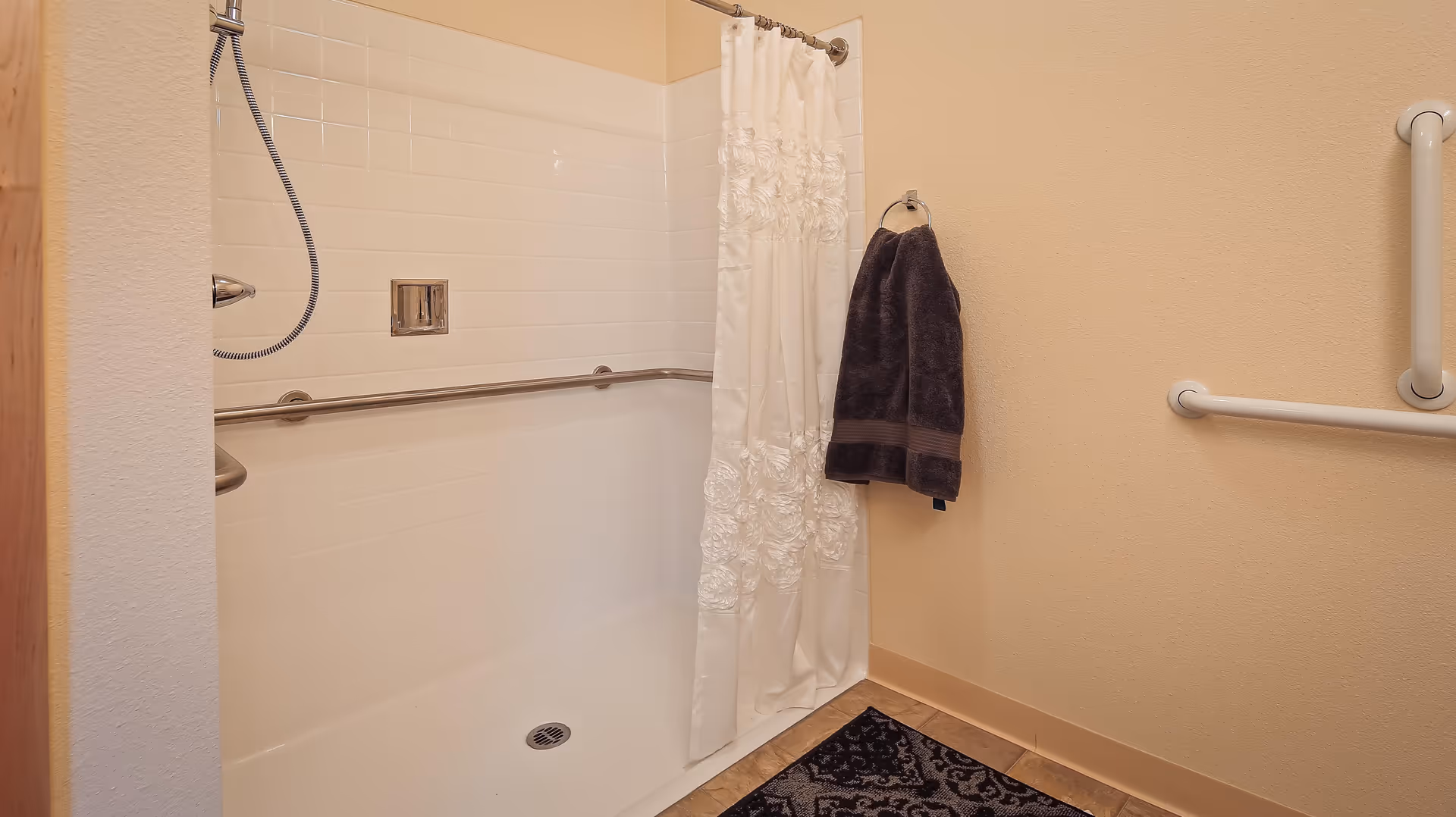 A bathroom shower area with white tiled walls and a white shower curtain with floral patterns. There are metal grab bars installed on the walls for support, a handheld showerhead, a dark towel hanging on a ring, and a patterned black and gray rug on the floor.